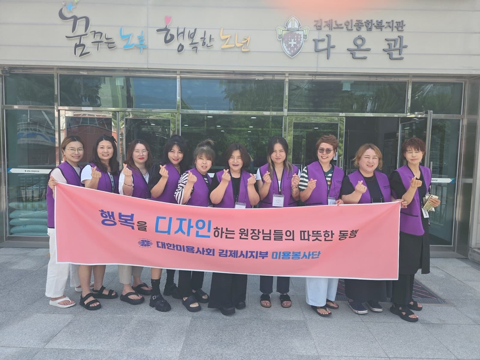 복지관 다온관 앞에서 기념 촬영한 미용봉사단 — 현수막에 '행복을 디자인하는 원장님들의 따뜻한 동행'이라 적혀 있다